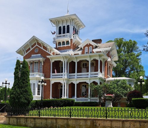 A grand Victorian-style mansion with a tower, surrounded by lush greenery and a decorative iron fence.