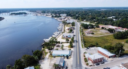 Aerial view of a riverfront town with boats, a road, and greenery, showcasing a mix of buildings and natural scenery.