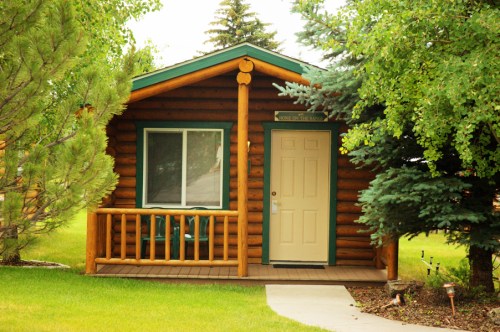 A small log cabin with a porch, surrounded by green trees and grass, featuring a front door and a large window.