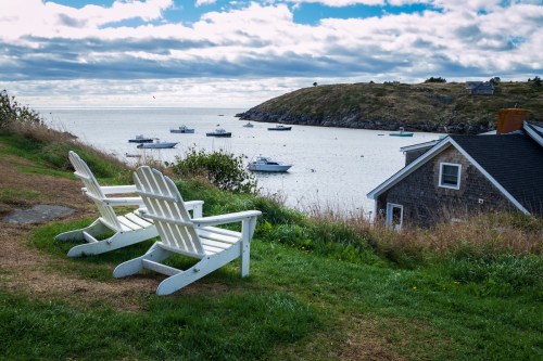 Two white Adirondack chairs overlook a calm bay with boats and a grassy hillside under a cloudy sky.