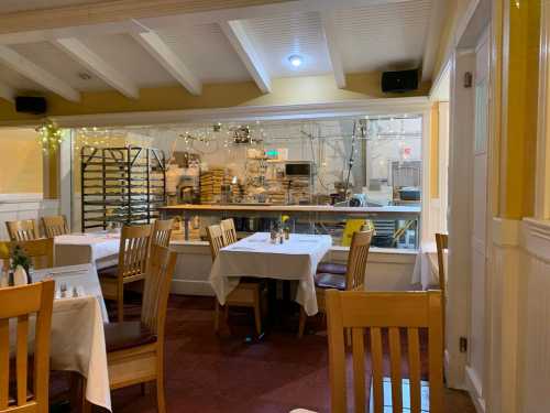 Cozy restaurant interior with white tablecloths, wooden chairs, and a view of a kitchen area with equipment and lights.