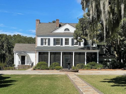 A charming two-story house with a porch, surrounded by greenery and trees, under a clear blue sky.