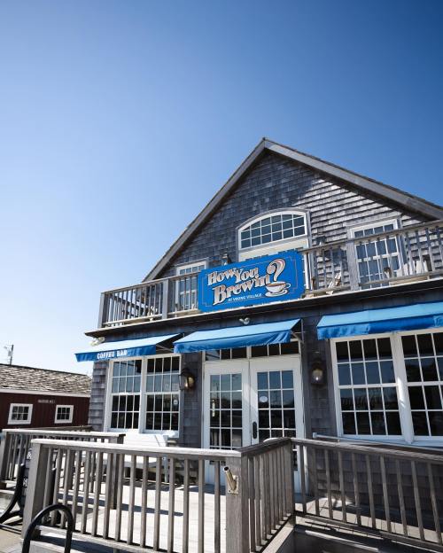 A charming coffee bar with a blue sign reading "How You Brewin'?" and a wooden deck under a clear blue sky.