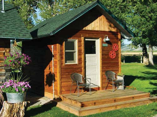 A cozy wooden cabin with a green roof, featuring a porch, two chairs, and flower pots in a sunny outdoor setting.