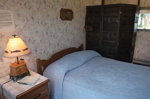 Cozy bedroom with a blue quilted bedspread, wooden furniture, a lamp, and a folding screen in a floral wallpaper setting.