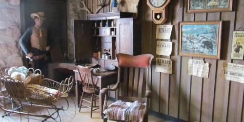 A vintage room with a wooden desk, chair, and wall decor, featuring a mannequin and an old-fashioned baby carriage.
