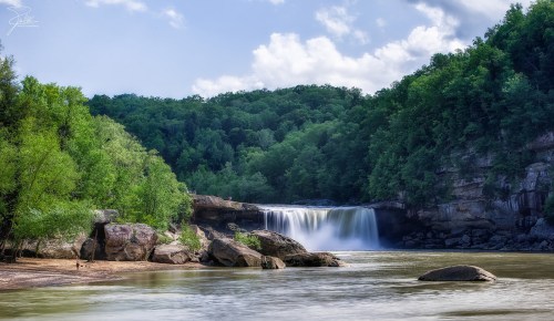 A serene waterfall cascades into a calm river, surrounded by lush green trees and rocky formations under a blue sky.