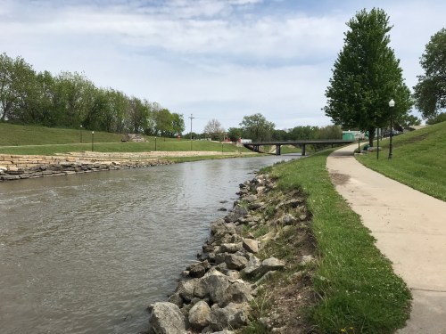 A calm river flows alongside a paved path, with greenery and a bridge in the background under a cloudy sky.
