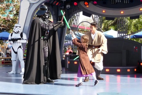 A child in a Jedi costume battles Darth Vader with a lightsaber, while a Jedi mentor watches in a themed performance.