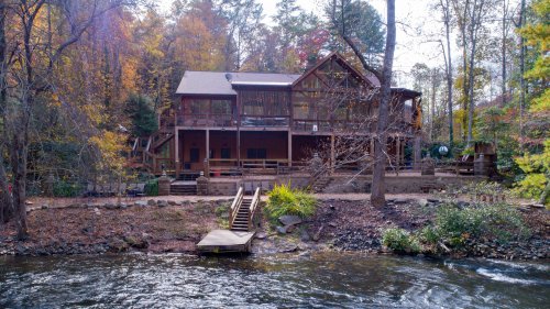 A rustic cabin by a river, surrounded by autumn trees, with a wooden deck and stairs leading to the water.
