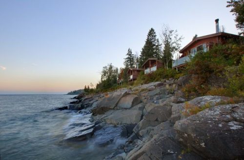 A serene lakeside view with wooden cabins nestled among trees, rocky shoreline, and calm water at sunset.