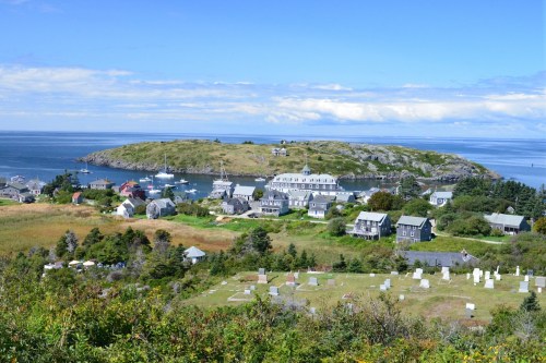 A scenic view of a coastal village with houses, a harbor, and a cemetery, surrounded by lush greenery and blue skies.