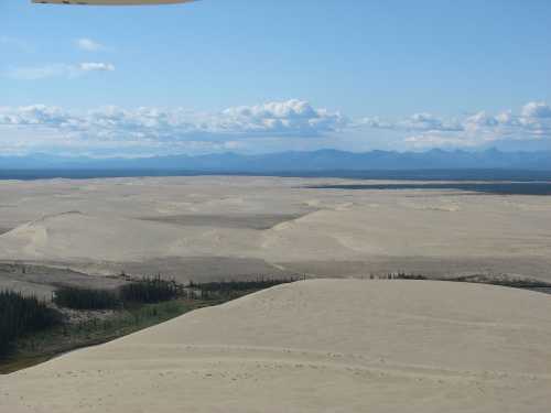 A vast desert landscape with rolling sand dunes, sparse vegetation, and distant mountains under a blue sky with clouds.