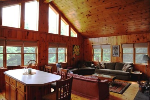 Cozy wooden living room with large windows, a dining table, and comfortable seating. Natural light fills the space.