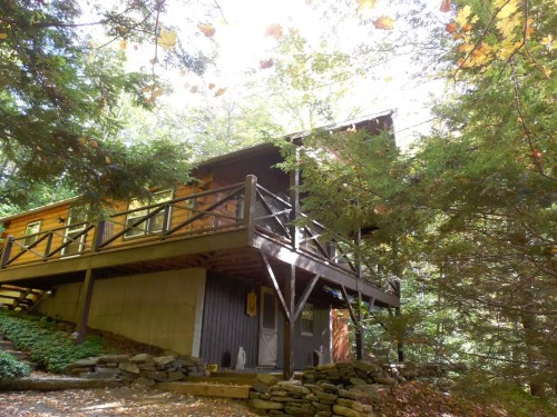 A cozy wooden cabin nestled among trees, featuring a balcony and stone pathway in a serene forest setting.