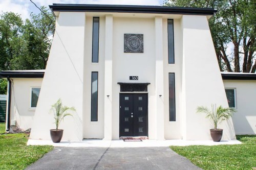 Modern building with a unique triangular design, featuring a black door and large windows, surrounded by greenery.