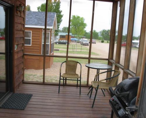 Screened porch with two chairs and a small table, overlooking a gravel area with parked cars and a cabin in the distance.
