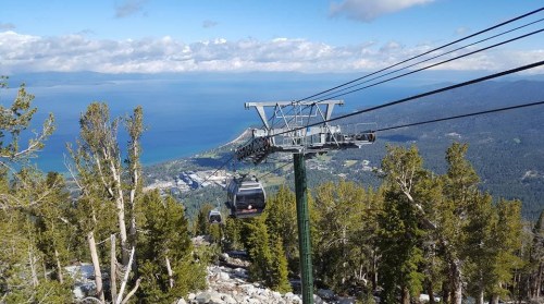 A scenic view of a gondola lift over a mountainous landscape, with a lake and clouds in the background.