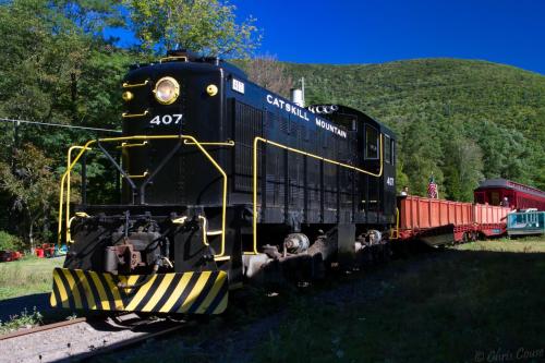 A black vintage locomotive with "Catskill Mountain" on its side, parked beside a red train car in a scenic green landscape.