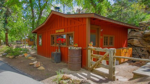 A charming red cabin surrounded by trees, featuring a wooden fence and stone pathway.