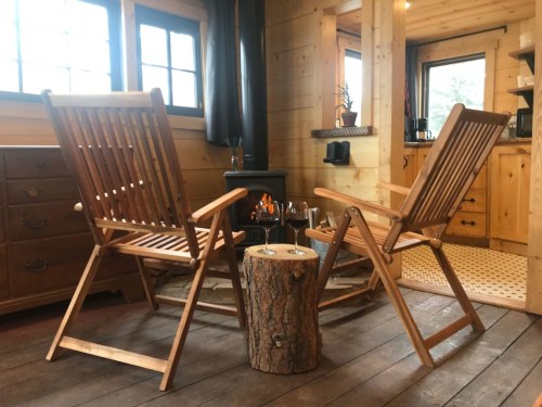 Two wooden chairs facing a small fireplace, with a log table and glasses nearby, in a cozy wooden interior.