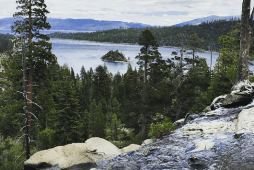 A scenic view of a lake surrounded by trees, with a small island in the distance and rocky terrain in the foreground.