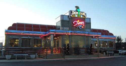 A retro-style diner with neon lights and glass block windows, featuring a colorful sign and outdoor seating.