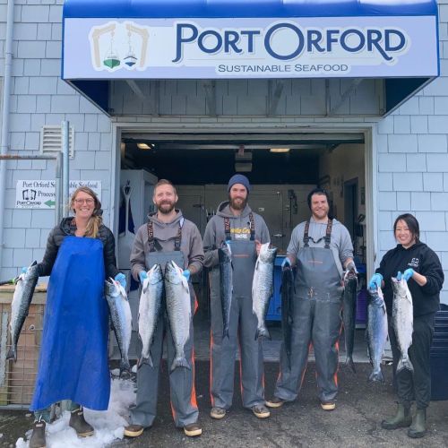 Five people in fishing gear hold large fish outside a seafood processing facility with a sign reading "Port Orford Sustainable Seafood."