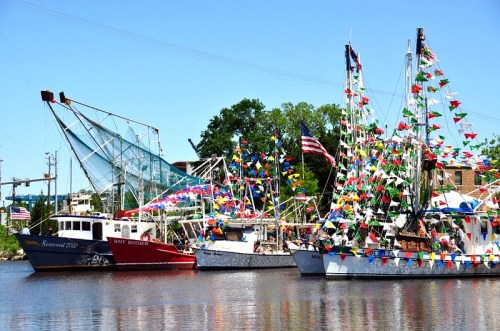Colorful fishing boats decorated with flags and American flags docked in a calm harbor on a sunny day.