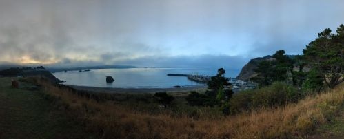A panoramic view of a calm bay surrounded by foggy hills and trees, with a small harbor in the distance.