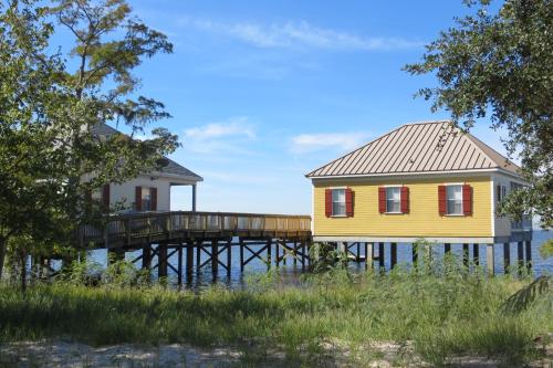 Two colorful houses on stilts by the water, connected by a wooden walkway, surrounded by greenery and blue skies.