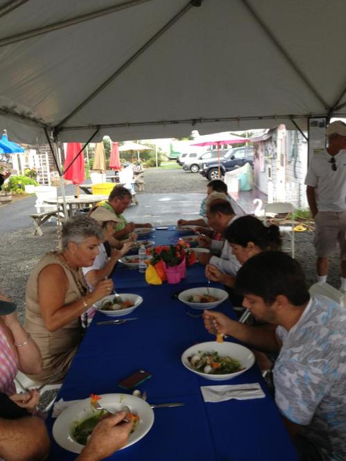A group of people dining under a tent, enjoying salads at a long table with colorful decorations.