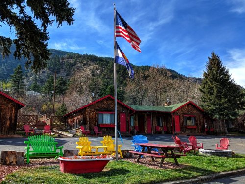 Colorful chairs surround a flower bed in front of rustic cabins, with flags waving against a backdrop of mountains and blue sky.