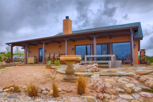 A modern adobe-style house with large windows, a stone patio, and a decorative fountain, set against a cloudy sky.