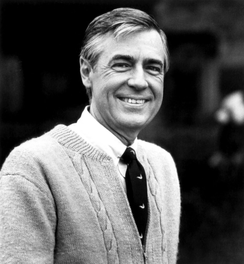A smiling man in a light sweater and tie, standing outdoors with a blurred background.