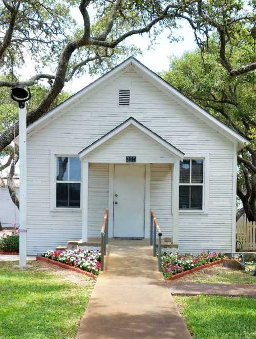 A small white house with a front porch, surrounded by colorful flowers and trees, on a sunny day.