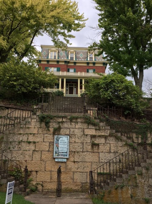 A large, historic brick house with green shutters, surrounded by trees, viewed from a stone staircase.