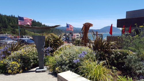 A scenic view featuring flags, sculptures, and vibrant flowers with a coastal backdrop and mountains in the distance.