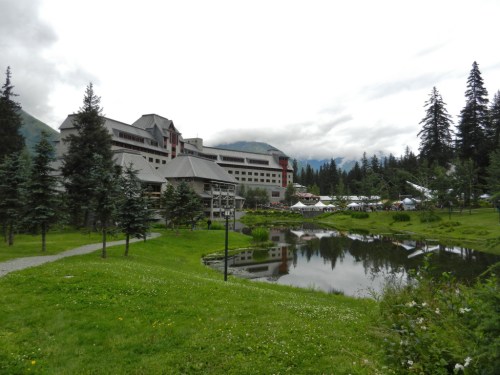 A scenic view of a hotel by a pond, surrounded by trees and mountains under a cloudy sky.