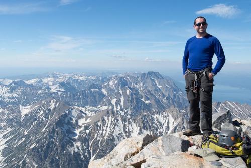 A climber stands on a mountain peak, wearing a blue shirt, with snow-capped mountains and a clear sky in the background.