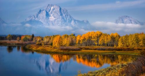 A serene landscape featuring mountains, autumn trees, and a reflective lake under a clear blue sky.