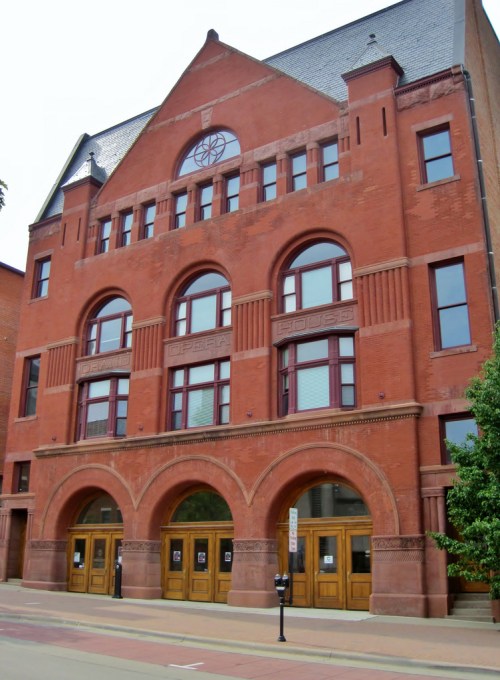 Historic red brick building with arched windows and doors, featuring decorative stonework and a triangular roof.