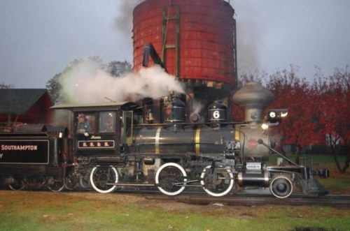A vintage steam locomotive with a red water tower, emitting steam in a scenic, autumn setting.