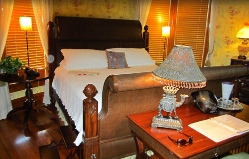 Cozy bedroom with a large bed, wooden furniture, a lamp, and decorative items, illuminated by warm light.