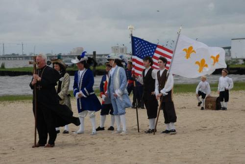 A group of people in historical costumes, including flags, walking along a beach with a cloudy sky in the background.