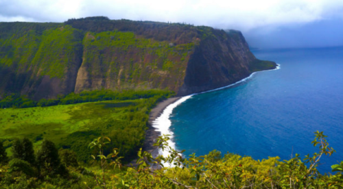 A scenic view of a lush green cliffside meeting a blue ocean, with a curved shoreline under a cloudy sky.