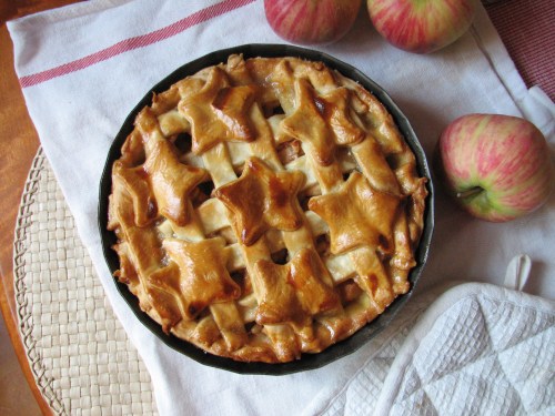 A freshly baked apple pie with a star-shaped lattice crust, surrounded by red apples on a white cloth.