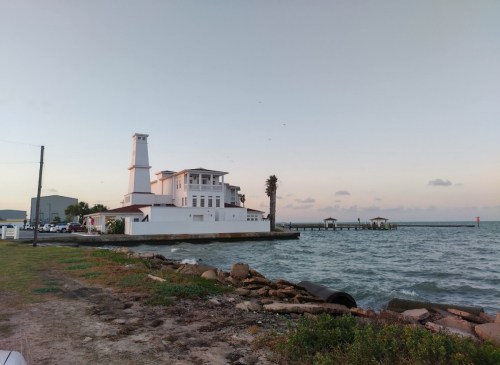 A coastal scene featuring a white building, palm trees, and a pier extending into the water at sunset.