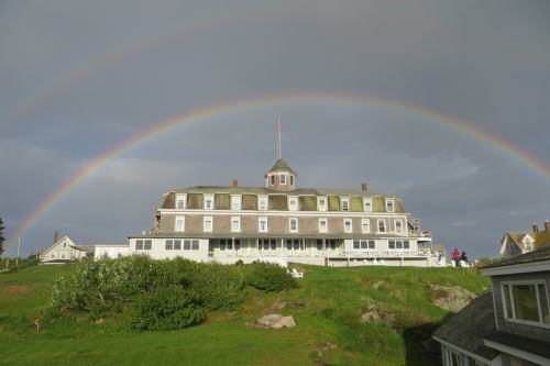 A large historic building on a green hill under a double rainbow against a cloudy sky.