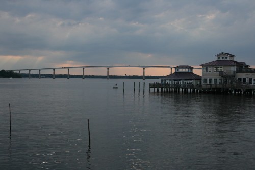 A calm body of water reflects a bridge and a waterfront building under a cloudy sky at dusk.